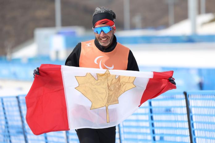 A male athlete wearing sunglasses and an Orange bib holds the Canadian flag